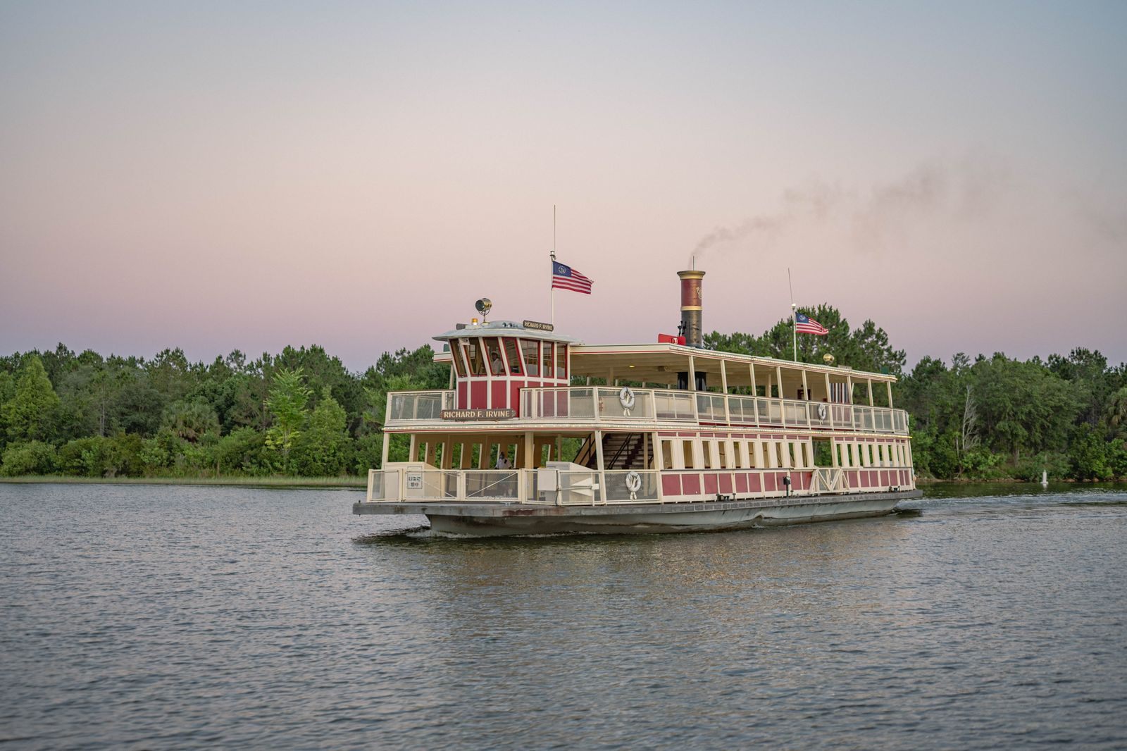 Cream-and-red riverboat with a single stack cruising past green trees at dusk