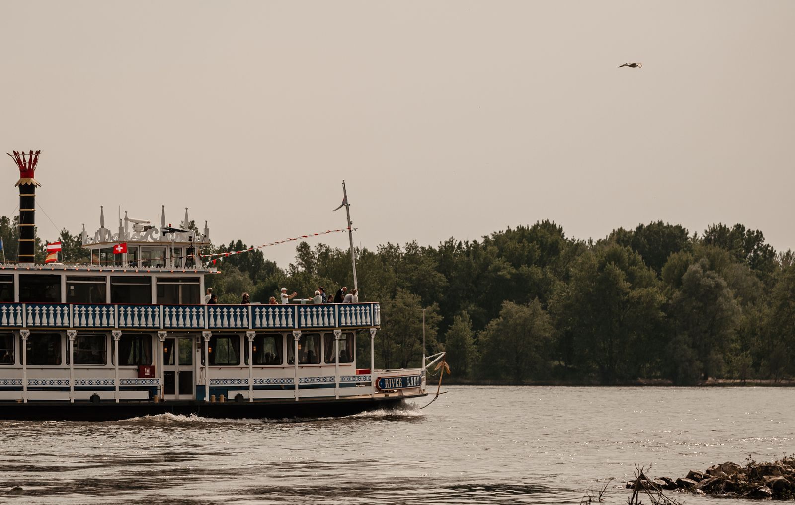 Riverboat with a tall red stack cruising past a tree-lined riverbank